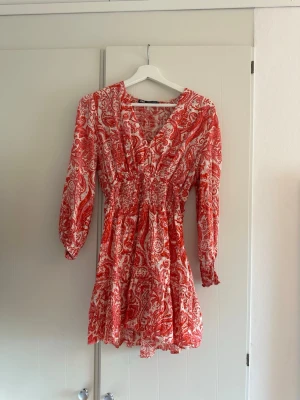 Röd och vit kortklänning från Zara - Supersöt kortklänning från Zara i rött och vitt med paisleyinspirerat mönster. Klänningen har v-ringning, långa ärmar med resår vid ärmslut och smock i midjan för en snygg passform. Perfekt för sommarens alla tillfällen!