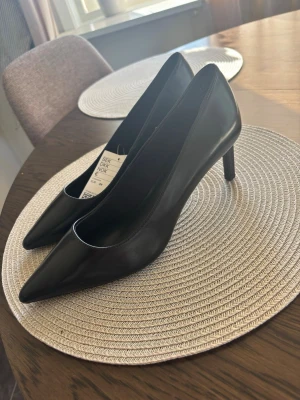 Svarta klassiska pumps från H&M - Stilrena svarta pumps från H&M med spetsig tå och smal klack. Skorna är tillverkade i ett slätt, glansigt material som ger en elegant look. Perfekta för dig som gillar en tidlös och chic stil.