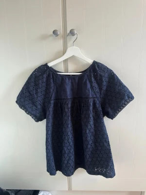 Mörkblå blus med broderade detaljer - Söt mörkblå blus från Next med korta ärmar och broderade hålmönster över hela tyget. Blusen har en lös passform och rund halsringning, perfekt för en avslappnad och trendig look. Tillverkad i mjuk bomull som känns skön mot huden.