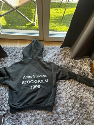  hoodie Acne Studios  - Snygg hoodie från Acne Studios med stor vit text på ryggen: 'Acne Studios STOCKHOLM 1996'. Klassisk passform med huva och ribbade muddar. Perfekt för dig som gillar stilrena och trendiga plagg med streetwear-vibe. 