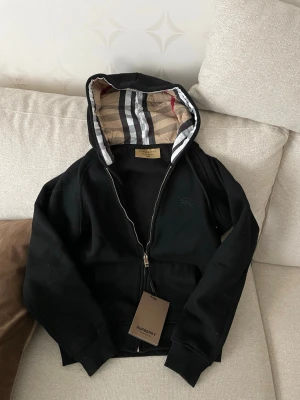 Svart hoodie från Burberry - Snygg svart hoodie från Burberry med dragkedja och klassiskt rutigt foder i huvan. Hoodien har fickor framtill och ribbade muddar vid ärmar och nederkant. Perfekt för dig som gillar stilrena plagg med exklusiva detaljer, den passar s också 