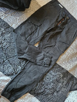 Svarta cargobyxor från Jack & Jones - Svarta cargobyxor från Jack & Jones med flera praktiska fickor på sidorna och resår vid bensluten. Byxorna har en avslappnad passform och är tillverkade i bomullsmix för skön känsla. Perfekta för en streetwear-look.