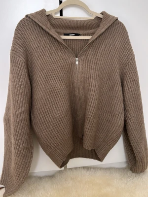 Beige stickad hoodie från BikBok - Mysig beige stickad hoodie från BikBok med dragkedja framtill. Tröjan har en avslappnad passform, huva och ribbstickade detaljer. Perfekt för lager-på-lager och chill dagar. Materialet är mjukt och ger en skön känsla.