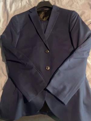 Blå kostym från Jack & Jones - Snygg blå kostym från Jack & Jones med klassisk kavaj och matchande byxor. Kavajen har två knappar och smal passform, perfekt för en stilren look. Kostymen är helfärgad och har en mjuk känsla i materialet. Storleken är 46/S 