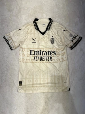 AC Milan bortatröja Puma M - Säljer en snygg AC Milan bortatröja från Puma i storlek M. Tröjan är cremevit med guldiga detaljer och svarta loggor, har korta ärmar och svart krage med mönster. Tillverkad i lätt och ventilerande material, perfekt för fotboll eller träning.