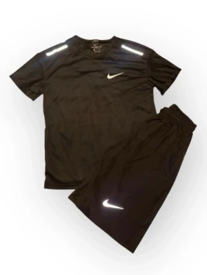 Svart träningsset från Nike - Säljer ett svart träningsset från Nike med t-shirt och shorts. Båda plaggen har vita Nike-loggor och reflekterande detaljer på axlar och ben. Setet är gjort i ett lätt och snabbtorkande material som passar perfekt för träning och sport.