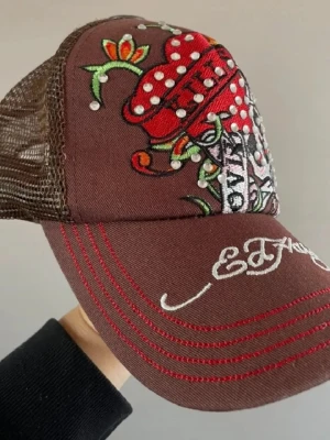 Ed Hardy keps - Snygg brun keps från Ed Hardy med broderat döskallemotiv, röda och vita detaljer samt nitar. Kepsen har mesh på sidorna och justerbar baksida. Märkeslogga broderad framtill på skärmen. Perfekt för dig som gillar streetstyle och unika accessoarer.