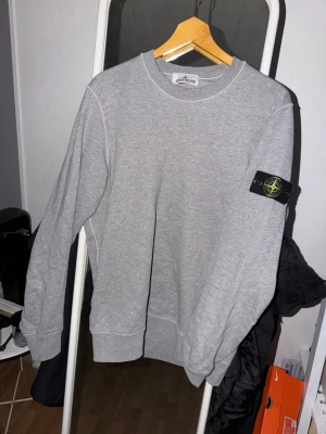 Grå sweatshirt från Stone Island - Snygg grå sweatshirt från Stone Island med klassisk rund hals och ikonisk logotyp-patch på vänster ärm. Tröjan har en clean look med ribbade muddar och är perfekt för en avslappnad streetwear-stil.