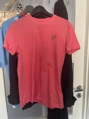 Rosa t-shirt från Lyle & Scott - Snygg rosa t-shirt från Lyle & Scott med klassisk rund hals och korta ärmar. På bröstet sitter den ikoniska gula loggan i form av en liten fågel. Perfekt för en avslappnad och trendig stil.