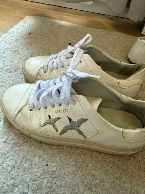 Vita sneakers från Axel Arigato - Snygga vita sneakers från Axel Arigato med broderade fåglar på sidan och klassiska vita skosnören. Skorna är i läder och har en stilren, minimalistisk design som passar till det mesta. Perfekta för dig som gillar clean och trendig stil.
