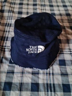 Mörkblå bucket hat The North Face - Snygg mörkblå bucket hat från The North Face med broderad logga framtill och logotyptext längs kanten. Klassisk form och ventilerande öljetter på sidorna. Perfekt accessoar för en avslappnad streetwear-look.