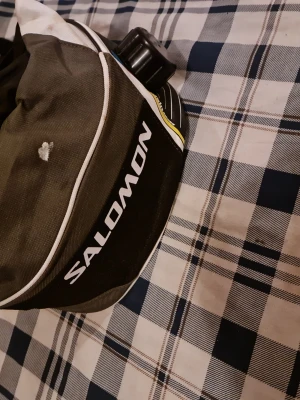 Salomon midjeväska med vätskebehållare - Sportig midjeväska från Salomon i svart och grått med vita detaljer och logga. Väskan har inbyggd vätskebehållare med skruvlock, perfekt för löpning eller träning. Tillverkad i slitstarkt syntetmaterial och har justerbart band.