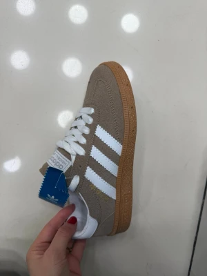 Adidas beige sneakers (finns i storlek 37-40) - Snygga Adidas sneakers i beige mocka med klassiska vita ränder och vit häl. Skorna har vit plös, vita skosnören och en brun gummisula. Perfekt för dig som gillar stilrena och tidlösa sneakers med retrovibbar.