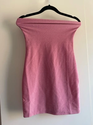 Rosa axelbandslös kort klänning - Trendig rosa axelbandslös kort klänning med glittrig finish. Klänningen har en tight passform och är perfekt för dig som vill sticka ut. Materialet känns mjukt och stretchigt, vilket gör den bekväm att bära hela kvällen. Köpt begagnat, endast provad. Har lite trådsläpp. Obs katt finns i hem!