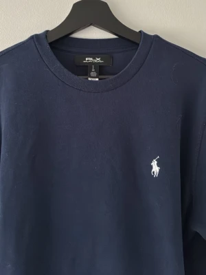 Mörkblå crewneck från Polo Ralph Lauren - Mörkblå sweatshirt från Polo Ralph Lauren med klassisk vit broderad logga på bröstet. Tröjan har rund halsringning, långa ärmar och ribbade muddar. Perfekt för en stilren och avslappnad look.