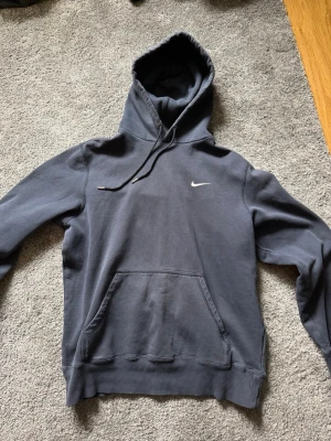 Mörkblå Nike hoodie med huva - Snygg mörkblå hoodie från Nike med klassisk vit swoosh-logga på bröstet. Hoodien har en stor känguruficka framtill, ribbade muddar och justerbar dragsko i huvan. Perfekt för en avslappnad och sportig stil.