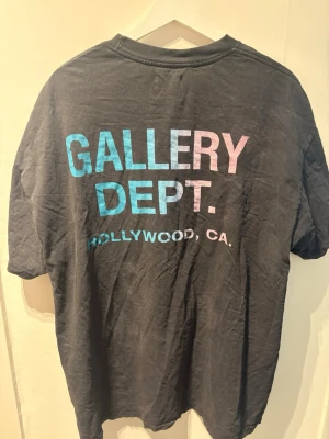 Svart Gallery Dept. t-shirt XL - Cool svart t-shirt från Gallery Dept. med färgglad text på ryggen i blått och rosa som säger 'GALLERY DEPT. HOLLYWOOD, CA.'. Klassisk passform och rund halsringning, perfekt för en avslappnad streetwear-look.