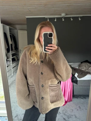 Beige teddyjacka med stora fickor - Mysig beige teddyjacka med knäppning framtill och två stora fickor i mockaliknande material. Jackan är grå brun. 