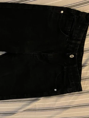 Svarta raka jeans - Säljer ett par svarta jeans med rak passform och klassisk femficksdesign. Jeansen har silverfärgade nitar och knapp, samt bälteshällor. Materialet är denim i bomull. Perfekta till en avslappnad och stilren look.
