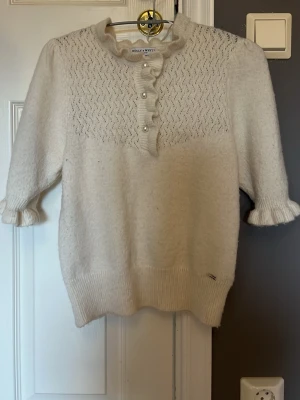 Vit stickad topp från Hm - Söt vit stickad topp från Hm med korta ärmar, volangkant vid halsen och pärlknappar framtill. Fint hålmönster upptill och ribbade muddar vid ärmslut och nederkant.