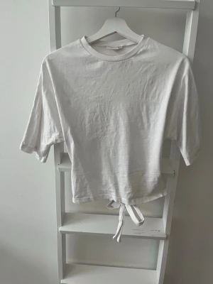 Vit topp med knyt - Säljer en vit topp i bomull med korta ärmar och knytband i ryggen. Enkel och clean design som passar perfekt till jeans eller kjol. Rund halsringning och mjukt material för en avslappnad look. Köparen står för eventuell frakt.
