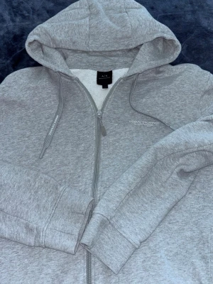 Grå zip-hoodie från Armani Exchange - Snygg gråmelerad hoodie från Armani Exchange med dragkedja framtill och justerbar huva. Diskret logga tryckt på bröstet och mjukt material som känns skönt mot huden. Perfekt för en chill och stilren look.