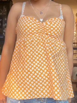 Gul blommig linnetopp - Supersöt gul linnetopp med vita små blommor och rynkdetalj vid bysten. Toppen har smala axelband och en luftig, lätt utsvängd passform som är perfekt för varma dagar. Perfekt att styla med jeans eller kjol för en fräsch sommarlook.