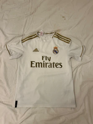 Real Madrid matchtröja Adidas vit - Säljer en vit Real Madrid matchtröja från Adidas med guldiga detaljer och klubbmärke broderat på bröstet. Tröjan har korta ärmar, rund hals och tre guldiga ränder på axlarna. Materialet är lätt och andas, perfekt för fotbollsträning eller match.