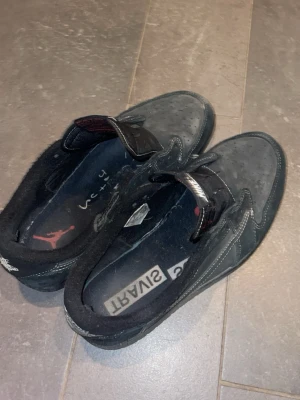 Svarta Nike Air Jordan 1 Low sneakers - Säljer ett par svarta Nike Air Jordan 1 Low sneakers med klassisk swoosh på sidan och broderad logga på hälen. Skorna har mockadetaljer, vita sömmar och kardborreband istället för vanliga snören. Perfekta för dig som gillar streetwear och vill ha något unikt.