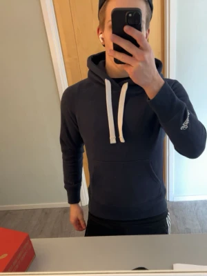 Russemerch hoodie  - Snygg mörkblå hoodie från russemerch med vit dragsko och broderad logga på ärmen. Klassisk känguruficka framtill och ribbade muddar. Använd 4-5 ggr. Säljer för den är för liten