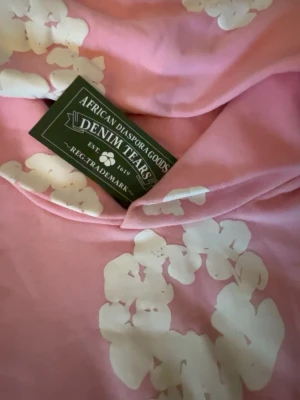 Rosa hoodie från Denim Tears - Säljes en rosa hoodie från Denim Tears med vita blommönster över hela tröjan. Hoodien har huva och långa ärmar, och är gjord i mjukt bomullsmaterial. Perfekt för dig som vill sticka ut med en unik och trendig look.