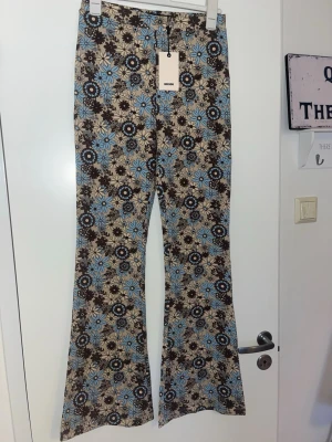 Mönstrade utsvängda byxor från Bik Bok - Säljer ett par retroinspirerade byxor från Bik Bok med utsvängda ben. De har ett beige, brunt och blått blommönster över hela byxan. Materialet är stretchigt och mjukt, perfekt för dig som gillar statement-plagg och vill sticka ut.