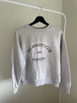 Grå sweatshirt NYC Athletics Club - Ljusgrå sweatshirt med tryck 'NYC ATHLETICS CLUB 1999 MANHATTAN NEW YORK' på bröstet. Tröjan har rund halsringning, ribbade muddar vid ärmslut och nederkant. Perfekt för en avslappnad streetwear-look.