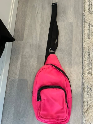 Rosa crossbody ryggsäck - Säljer en snygg rosa crossbody ryggsäck med svart justerbar rem och dragkedjor. Väskan har ett stort huvudfack och en mindre ficka framtill, perfekt för att bära det viktigaste. Tillverkad i slitstarkt syntetmaterial med enkel och modern design.