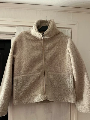 Beige teddyjacka med dragkedja - Mysig beige teddyjacka med hög krage och dragkedja framtill. Jackan har två stora fickor och är gjord i fluffigt, mjukt material som ger en avslappnad look. Perfekt för kyliga dagar och enkel att matcha med olika outfits.