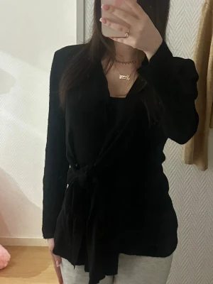 Svart omlottkofta med knytband - Snygg svart omlottkofta med långa ärmar och knytband i midjan. Koftan har en djup V-ringning och är tillverkad i ett mjukt, ribbat material som ger en skön passform. Perfekt att slänga över en topp för en stilren look.