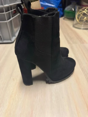 Svarta stövletter med klack - Säljer ett par svarta stövletter med chunky klack och grov sula. Skorna har dragkedja på insidan och är gjorda i mockaimitation. Perfekta för dig som vill ha en edgy look och gillar höga klackar. Passar till både jeans och kjol.