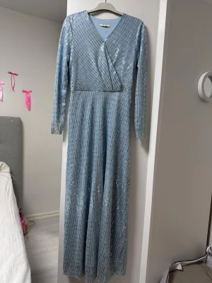 Ljusblå glittrig långklänning omlott - Säljer en ljusblå långklänning med glittriga paljetter och omlott överdel. Klänningen har v-ringning och långa ärmar, perfekt för speciella tillfällen. Materialet är troligtvis polyester och den har en elegant, svepande passform.