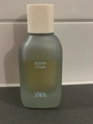 ZARA Sugar Foam parfym - ZARA Sugar Foam parfym i en stilren, frostad glasflaska med mjuka former och vit kork. Flaskan har en modern design med ljusgrå ton och tydlig logga framtill. Perfekt för dig som gillar trendiga och fräscha dofter.