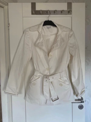 Vit trenchcoat med bälte från Vila - Stilren vit trenchcoat från Vila med markerad midja och tillhörande bälte. Jackan har stora knappar framtill, bred krage och två fickor med lock. Tillverkad i ett lätt tyg som passar perfekt till vår och sommar. Klassisk och tidlös design.