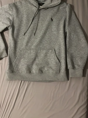 Grå hoodie från Polo Ralph Lauren - Snygg grå hoodie från Polo Ralph Lauren med klassisk huva och dragsnören. Har en känguruficka framtill och den ikoniska lilla svarta loggan broderad på bröstet. Tillverkad i mjukt bomullsmaterial och har ribbade muddar vid ärmslut och nederkant.