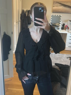 Svart snygg volangblus med v-ringning - Snygg svart blus i transparent material med volangdetaljer längs v-ringningen och ärmsluten. Blusen har långa ärmar och en lätt utsvängd passform nedtill. Perfekt både till vardags och finare tillfällen! Storlek xs, endast använd 1 gång så väldigt bra skick💕