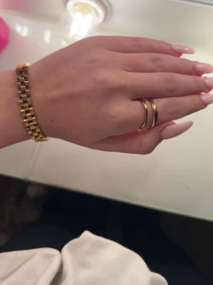 Dubbel guldfärgad ring i metall - Säljer en snygg dubbelring i guldfärgad metall med modern, rundad design. Ringen har en unik form där två ringar sitter ihop och ger ett statement-intryck. Perfekt för dig som gillar stilrena accessoarer med en twist.