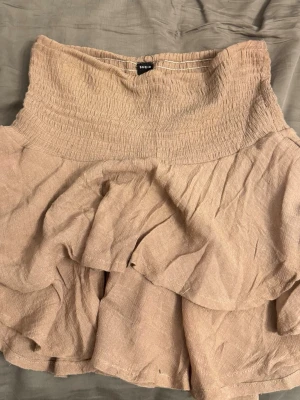 Brun volangkjol  - Säljer en  kortkjol med dubbla volanglager. Kjolen har en luftig och lätt struktur, perfekt för varmare dagar. Enkel att matcha med olika toppar för en trendig look. Kan även användas som topp.