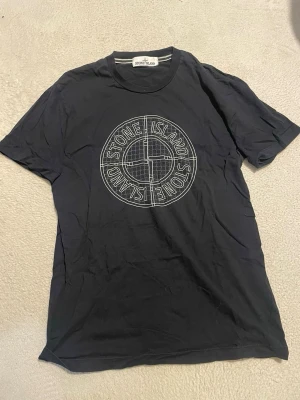 Svart Stone Island t-shirt - Snygg svart t-shirt från Stone Island med stort vitt logotryck på bröstet. Klassisk rund hals och korta ärmar. Perfekt för dig som gillar streetwear och vill ha en enkel men stilren look.
