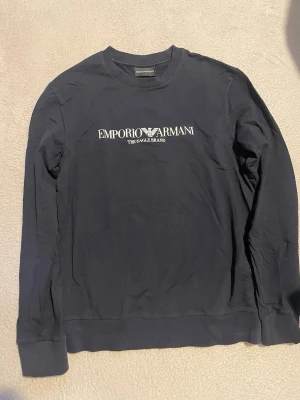 Marinblå sweatshirt från Emporio Armani - Snygg marinblå sweatshirt från Emporio Armani med vitt tryck och logga på bröstet. Tröjan har rund halsringning och långa ärmar. Perfekt för en stilren och avslappnad look.