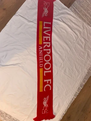 Liverpool FC röd halsduk - Röd halsduk med Liverpool FC-logga och texten 'LIVERPOOL FC ANFIELD' i vitt och gult. Halsduken har fransar i ändarna och är perfekt för att visa ditt stöd för laget. Klassisk design med klubbens emblem på båda sidor.