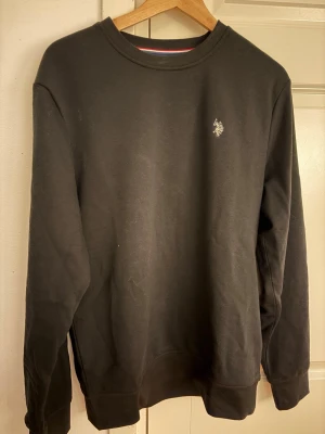 Svart sweatshirt från U.S. Polo Assn. - Klassisk svart sweatshirt med rund hals från U.S. Polo Assn. Tröjan har en liten broderad logga på bröstet och ribbade muddar vid ärmslut och nederkant. Perfekt för en avslappnad och stilren look. Knappast använd!
