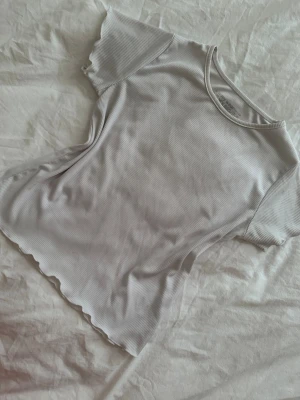 Vit ribbad t-shirt med vågig kant - Säljer en vit ribbad t-shirt med korta ärmar och vågig kant nertill. T-shirten har en rund halsringning och är gjord i ett mjukt, stretchigt material som sitter snyggt på kroppen. Perfekt till jeans eller kjol för en enkel men trendig look.