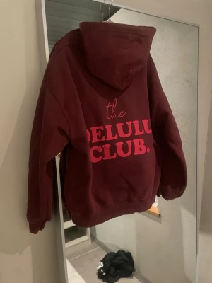 Vinröd hoodie med tryck DELULU CLUB - Snygg vinröd hoodie med stor huva och trycket 'the DELULU CLUB.' i rosa på ryggen. Oversized passform och mjukt material, perfekt för en chill och trendig look. Slutsåld 